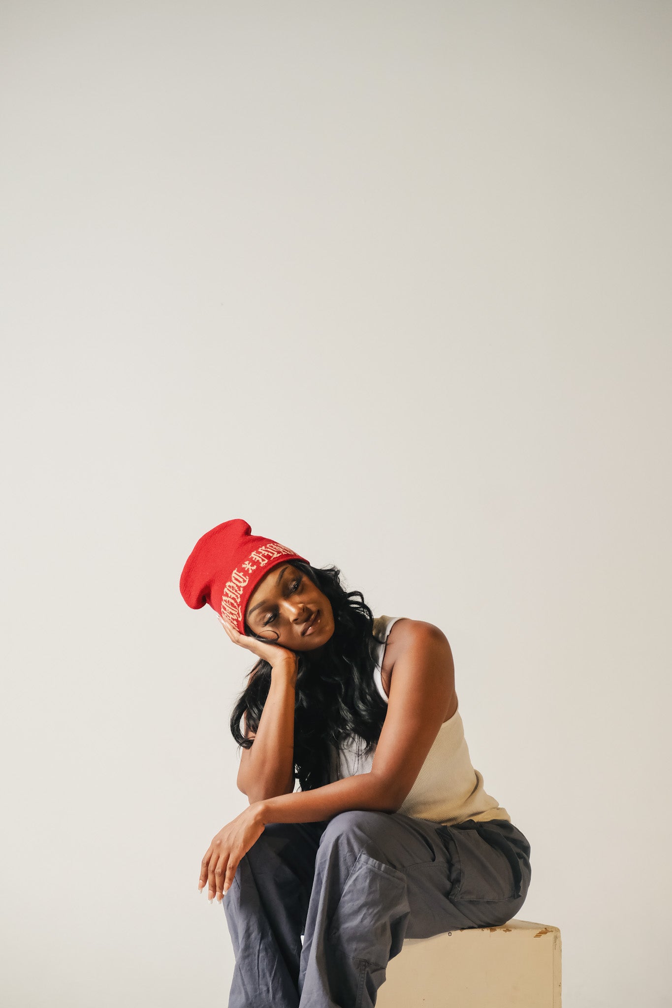 Model wearing a red beanie and sitting on a box against a plain background