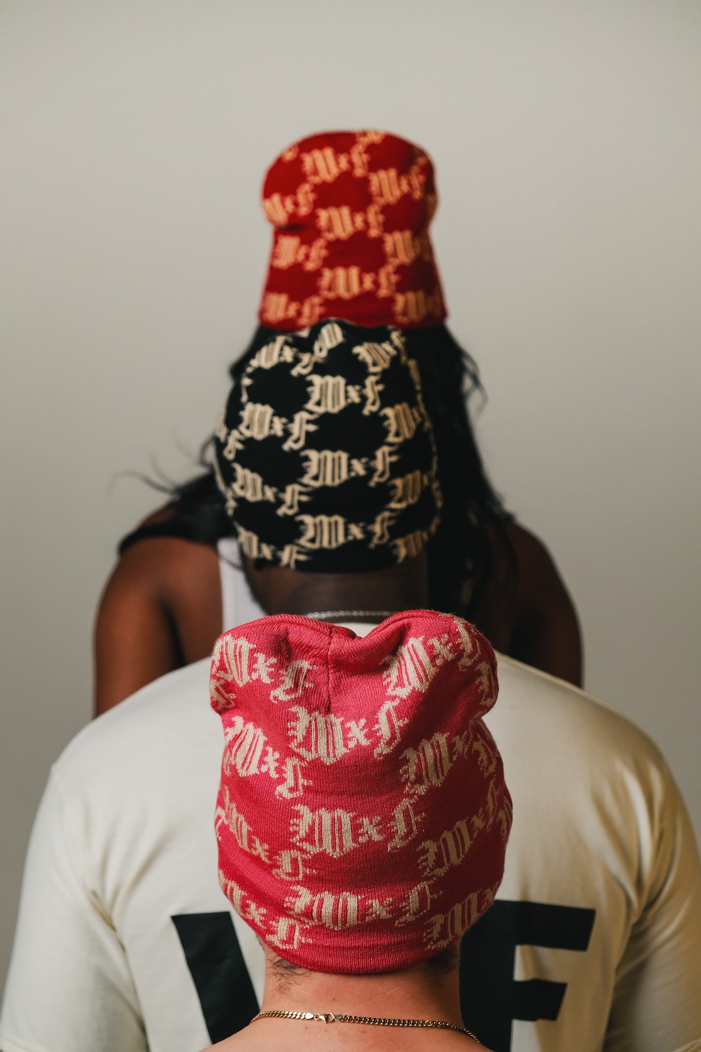 Three models wearing red, black, and pink beanies with monogram logo, facing away from the camera.